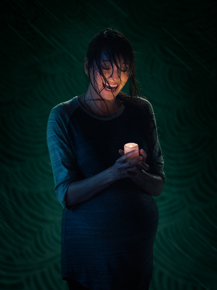 A photo for Woolly Mammoth used for the promotion of Baby Screams Miracle featuring a pregnant woman in the dark. She is soaked in her dress and holds a lit candle. She appears to be either laughing or crying.